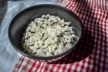 white parboiled black rice in a dark cup on a plate on a background of red-white cloth towels under the light of the sun with shadow on the table