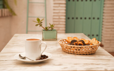 cup of coffee on marble table, with pastries in wicker basket and decorative plant, and background of Mediterranean house