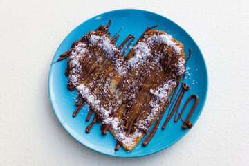Heart shape pancake with chocolate cream and grated coconut on blue plate and white background. Delicious sweet dessert
