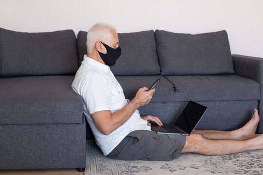 Online Job Concept. Serious Elderly Man In Medical Mask Working At Home On His Laptop Computer And Using Smartphone