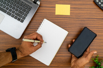 Writing on a notepad while holding a mobile phone working in an office environment. A lap top, a mobile, calculator and plant are also on display on this brown striped working table.