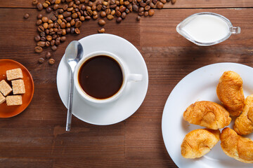 A cup of coffee on the table next to sugar and cream in a jug and a croissant.
