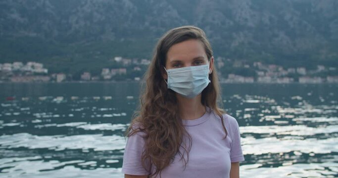 A young woman tourist with curly hair and a protective mask stands on a wooden pier. Mountain and bay views. Woman portrait close-up. High quality 4k footage