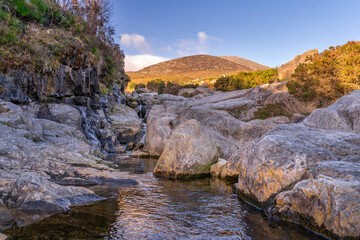 Mountain stream flowing between rocks with mountain peak of Slieve Donard, highlighted by sunlight in background, Mourne Mountains, Northern Ireland