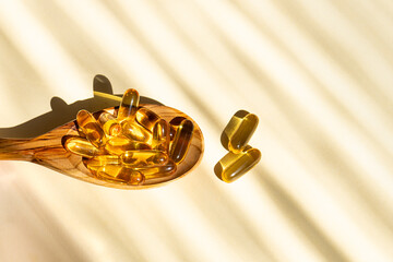 Cod liver oil capsules with vitamin D in the wooden spoon on light beige background with hard shadows. Health care concept.