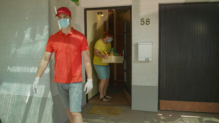 Courier wearing a surgical mask walks away after delivering groceries to lady.