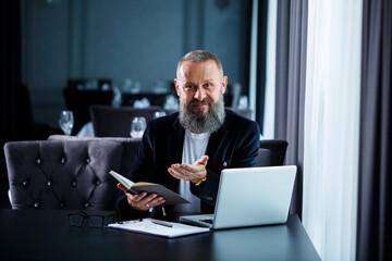 An adult successful male businessman is working on a new project and looks at the growth charts in a notebook. Sits at the table by the large window. Looks at a notebook and smiles