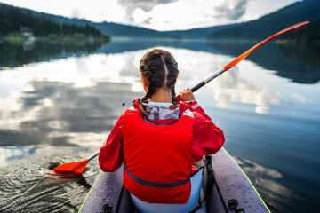 Caucasian woman in a kayak enjoys the clear view in front of her, with pine forest reflected in the beautiful and calm lake on a beautiful day of wild adventure. Hobby and leisure activity in nature.