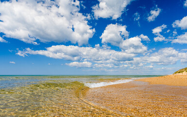 Seascape. Transparent clear sea on a sandy beach. A small wave washes the beach. In the background there is a blue sky with clouds.