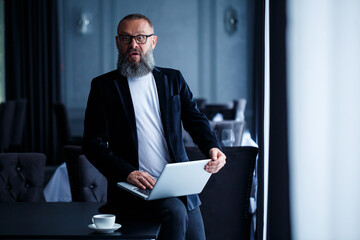 An adult businessman with a beard in glasses sits with a laptop on his lap and works. The director makes a schedule for the workflow