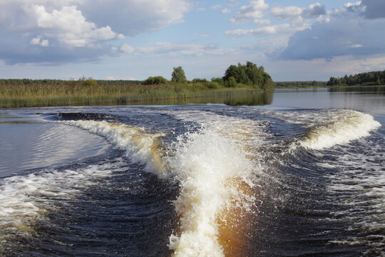 View from stern of fast floating motor boat on stern wake trail on river water at summer day on forestry banks and blue sky with white clouds background, natural riverscape view