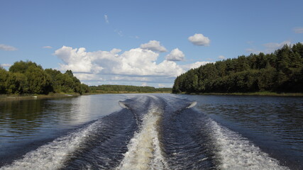 Beautiful view from the stern of a motor boat with an outboard motor on the transom Wake, floating on the calm water of the Volga river on a Sunny summer day on forest on shore and blue sky background