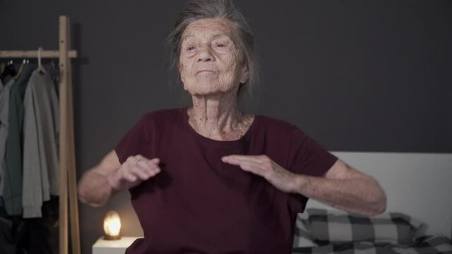Very old age woman with gray hair and deep wrinkles does physical exercises at home. An elderly female is warming up in a small apartment. Happy senior exercising fitness within doors during covid 19