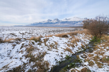 Snow-covered field with withered grass and bare bushes, the river flows against the backdrop of beautiful mountains