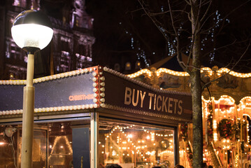 View of the ticket booth next to the glowing vintage carousel as part of the attractions in Princes Street Gardens, Edinburgh, UK, with a tree on the foreground.