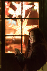 An Asian girls smiles and has her face lit up while using her phone in Princes Street Gardens, Edinburgh, UK, as she stands in front of a window with Christmas lights.