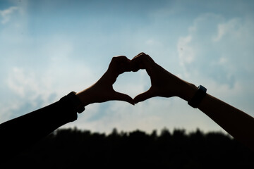 close up male and female hands making a heart shape silhouette in the sky.