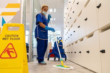 A cleaner with a mask on her face cleans the floor with the mop.Caution wet floor sign close up - obrazy, fototapety, plakaty