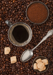 Cup of black coffee with ground coffee and cane sugar with silver spoon inside fresh coffee beans background.