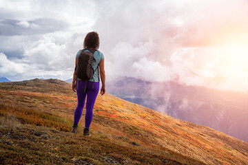 Fototapeta premium Adventurous Girl Hiking up the Nares Mountain during a cloudy and sunny evening. Taken at Carcross, near Whitehorse, Yukon, Canada.