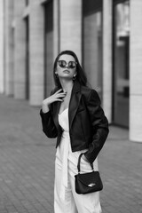 Fototapeta premium Young stylish woman in white overall, sneakers, black leather jacket and sunglasses holding small handbag and standing at city street. Fashion style outdoor portrait