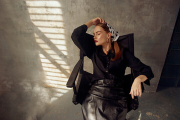 Stylish redhead sexy woman in black blouse, leather skirt and polka dot kerchief sitting at high...
