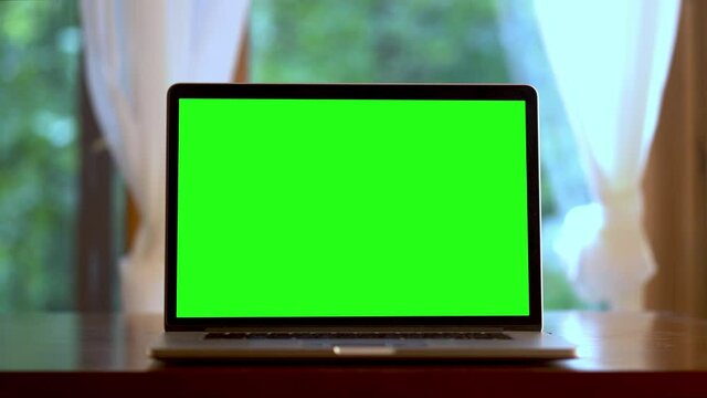 Fast dolly push in to laptop computer with green screen on a table in front of windows with curtains. Concept for remote virtual distance learning