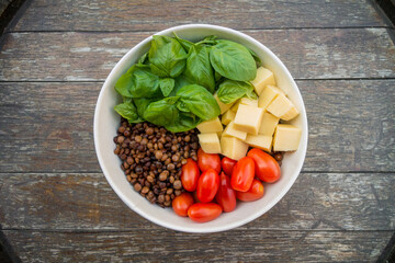 Top view of homemade vegetarian healthy salad. Colorful food, red cherry tomatoes, green basil, lentils and cheese in a white bowl on rustic wooden table.