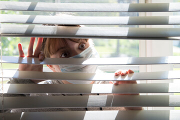 A young woman with face mask looks out from behind blinds 