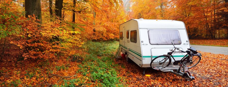 Caravan trailer with a bicycle parked in a golden beech tree forest. Colorful red, orange and yellow leaves on the ground. Panoramic autumn landscape. Leisure activity in Heidelberg, Germany