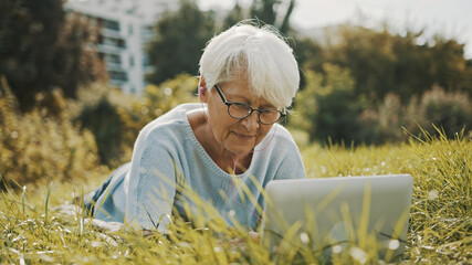 old gray haired woman using laptop in the nature while lying on the grass. High quality photo