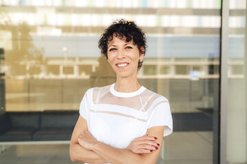 Portrait of smiling mature business woman standing arms crossed against glass outside office