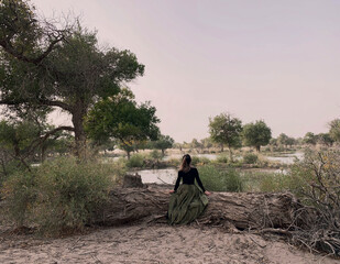 Girl sitting on trunk in desert Oasis with Populus euphratica forest and lake. Desert Conceptual Background. Lonely Tree In Surrealistic Sandy Environment fashion tourism travel concept Inner Mongolia
