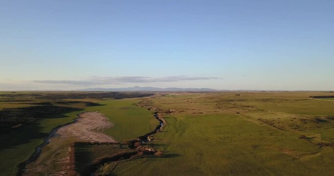 Aerial view of green rural field at sunset with stream going through it. Small mountains in the background.