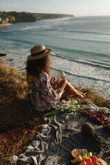 Picnic on the beach. Fruit table. Curly hair caucasian woman sit on the blanket on sea cliff and eat fruits