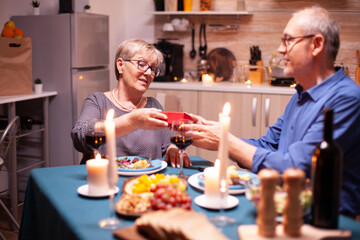 Elderly husband giving wife gift box during dinner. Happy cheerful elderly couple dining together at home, enjoying the meal, celebrating their anniversary, surprise holiday