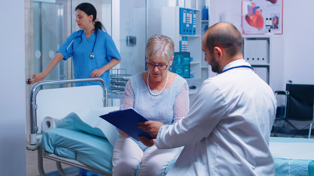 Senior Retired Woman Signing For Medical Release In Modern Private Clinic Sitting On Hospital Bed, Nurse Working In Background. Healthcare Medicine Consultation Doctor Patient