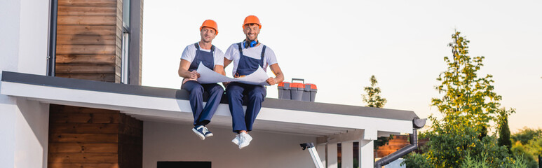 Panoramic shot of builders in uniform holding blueprint near toolbox on roof of building