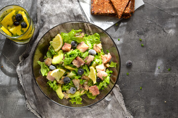 Salad with lettuce, fish, blueberries, lemon and avocado. Healthy lunch or brunch. Dark background. Linen gray napkin. Salad in a black glass plate. Backlight. Fish bowl with avocado. Top view.