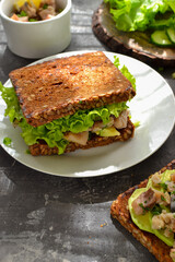 Dark bread sandwich with seeds. Sandwich with salad, avocado and fish in a white plate. Healthy lunch or brunch. Dark background. Breakfast at the cafe. Backlight. Vertical view