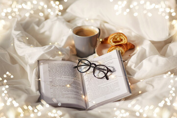 morning, hygge and breakfast concept - croissants, cup of coffee, book and glasses in bed at home