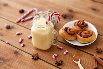 christmas and seasonal drinks concept - eggnog in glass mug with candy cane decoration, cinnamon buns and aromatic spices on wooden background