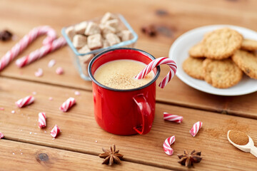 christmas and seasonal drinks concept - red cup of eggnog with candy cane, oatmeal cookies, fir tree branches and sugar on wooden background