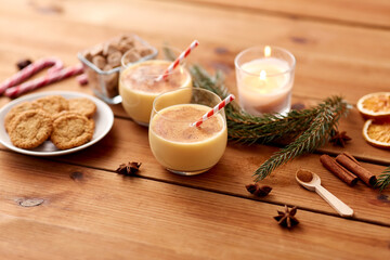 christmas and seasonal drinks concept - glasses of eggnog with oatmeal cookies, candy canes, sugar, fir tree branches and candle burning on wooden background