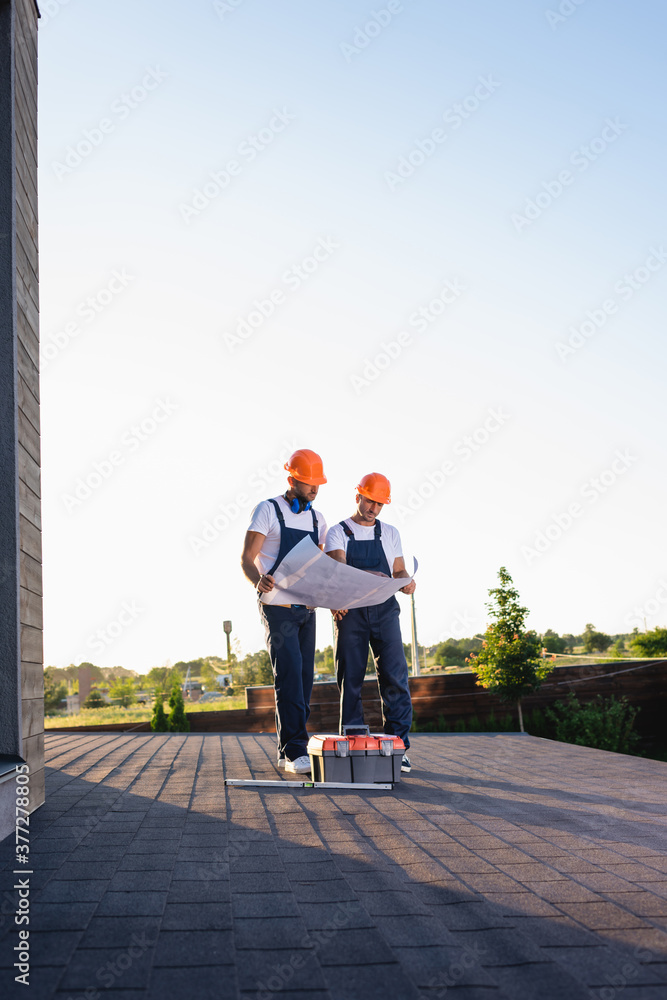 Wall mural builders with blueprint standing near toolbox on roof of building