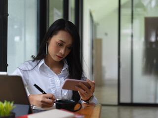 Female freelancer looking information on smartphone and write it down