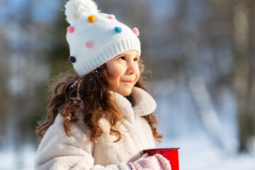 childhood, christmas and season concept - happy little girl with cup of hot tea in winter park