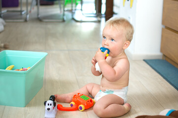 Ten month baby boy sitting on floor and playing with toy. Little kid in nappy at home. Baby care concept, banner copy space