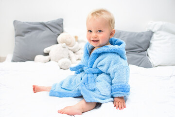 Ten month blond baby boy sitting on bed in blue bathrobe and smiling.White pillows in background. Baby care concept, banner copy space