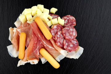 Meat and cheese snacking selection with prosciutto ham, salami and grissini bread sticks on a slate stone background